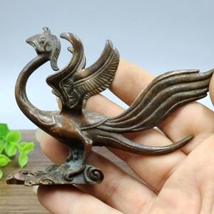 Pode incluir: Uma escultura de fénix de cor bronze com asas detalhadas e um pescoço longo e curvo. O pássaro está empoleirado numa base decorativa. A escultura é segurada na mão, mostrando o seu tamanho e design intrincado.