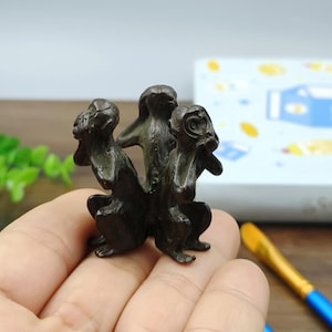 May include: A bronze-colored sculpture of three monkeys in a classic "see no evil, hear no evil, speak no evil" pose. The monkeys are clustered together, with one covering its eyes, another its ears, and the third its mouth. The sculpture is held in a hand.