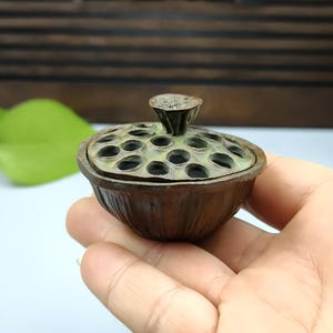 以下が含まれることがあります： 蓮の実の鞘の形をした、ブロンズ色の香炉。蓋には複数の円形の穴と、小さなテクスチャード加工のハンドルが付いています。香炉は手で持たれており、背景にはぼやけた緑の葉があります。
