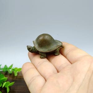 Op de afbeelding: Een kleine, bronzen schildpadfiguur. De schildpad heeft een afgerond schild en gedetailleerde poten en kop. Het beeldje wordt in een hand gehouden, wat de grootte en het vakmanschap laat zien. De algehele esthetiek is antiek of vintage.