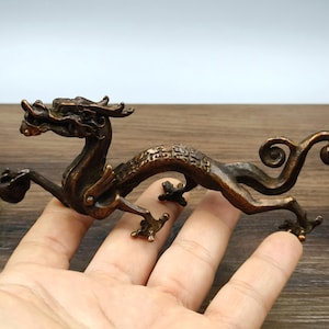 Op de afbeelding: Een bronzen drakenbeeldje met ingewikkelde details. De draak wordt afgebeeld in een dynamische pose, met zijn lichaam gebogen en zijn poten uitgestrekt. Het beeldje heeft een getextureerd oppervlak en een gedetailleerde kop.