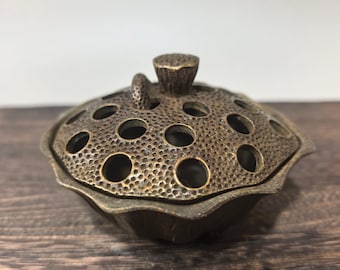 May include: A bronze-colored incense burner with a lotus flower design. The lid features multiple circular holes and a small, textured knob. The base has a petal-like shape, and the overall design is detailed and ornate.