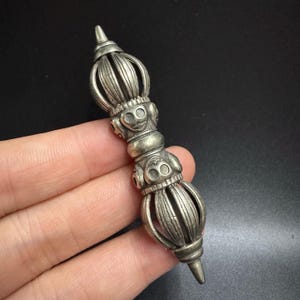 May include: A silver-coloured, ornate vajra, a ritual object with a symmetrical design. The vajra features a central bulbous section with faces and a pointed end. The object is held in a hand against a dark background.