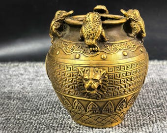 Puede incluir: Un recipiente de metal dorado y ornamentado, con cuerpo redondo y tapa. El recipiente está decorado con relieves de criaturas míticas y patrones geométricos. Tres criaturas están posadas en el borde, y una cabeza de león aparece en el frente.