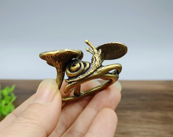 May include: A bronze-colored ring featuring a snail and mushroom design. The snail has detailed antennae and a shell, and is positioned near a mushroom. The ring is held by a hand.