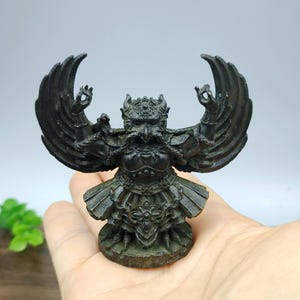 Può includere: Una statua in bronzo scuro di una creatura mitica simile a un uccello con le ali spiegate. La figura ha un volto dettagliato, un copricapo ornato e un indumento a forma di gonna. La statua è su una base rotonda.