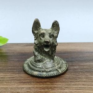 以下が含まれることがあります： 開いた口と詳細な毛並みを持つ、ブロンズ製の犬の置物。犬の頭は、装飾的で質感のある縁取りのある円形の台座に取り付けられています。置物は風化した外観をしています。
