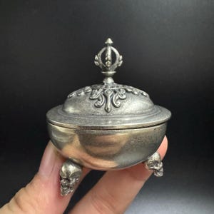 May include: A small, silver-coloured container with a decorative lid and three skull-shaped feet. The lid is adorned with ornate patterns and a stylized finial. The container appears to be made of metal and has a vintage or antique aesthetic.