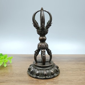 May include: A bronze-coloured Vajra, a ritual object, with intricate details. The top features a central point with curved elements and ornate carvings. The base is a lotus flower design. The object is approximately 15 cm tall.