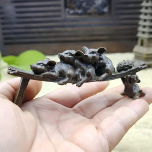 May include: A bronze-colored sculpture of four pigs resting on a wooden bench. A small pig supports a tray with a bundle of leaves. The sculpture is held in a hand, with a blurred background.