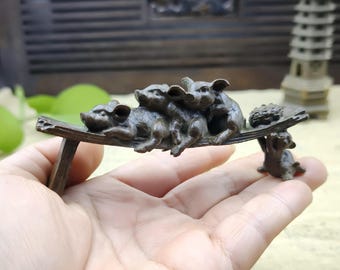 May include: A bronze-colored sculpture of four pigs resting on a wooden bench. A small pig supports a tray with a bundle of leaves. The sculpture is held in a hand, with a blurred background.
