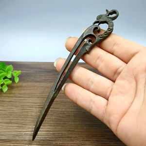 Puede incluir: Horquilla de bronce con diseño de dragón. La horquilla presenta un diseño dividido con una cabeza de dragón decorativa en la parte superior. La horquilla se sostiene en una mano, mostrando los intrincados detalles y la pátina envejecida del metal.
