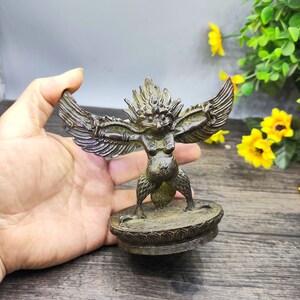 Pode incluir: Estátua de bronze de uma criatura mítica com asas abertas. A figura tem cabeça de pássaro, tronco humano e assenta numa base circular decorativa. A estátua é segurada na mão, com flores amarelas e folhagem verde ao fundo.