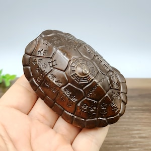 May include: A bronze-colored turtle figurine with a geometric shell pattern and engraved characters. The center of the shell features a yin-yang symbol. The figurine is held in a hand.