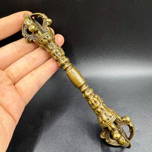 May include: A bronze-coloured Vajra, a ritual object with ornate detailing. The handle features sculpted faces and intricate patterns, with a three-pronged top. The object is held in a hand against a dark background.