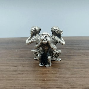 Puede incluir: Escultura plateada de los tres monos sabios: no ver el mal, no oír el mal, no hablar el mal. Los monos están sentados con las manos cubriendo los ojos, las orejas y la boca. La escultura está sobre una superficie de madera.