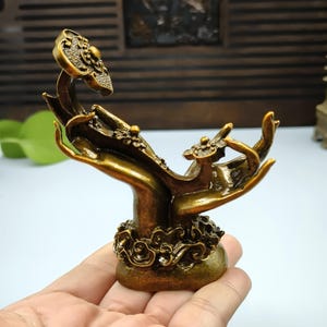 Puede incluir: Una escultura de color bronce que representa dos manos sosteniendo una flor. Las manos están adornadas con diseños detallados y descansan sobre una base con motivos florales. La flor presenta un diseño circular con una pequeña esfera dorada en el centro.