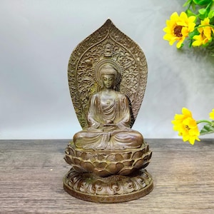 May include: A bronze-colored Buddha statue seated in a meditative pose on a lotus flower base. The statue has an ornate, patterned halo behind the head. The details include a serene facial expression and intricate carvings. The statue is a representation of Buddhist art.
