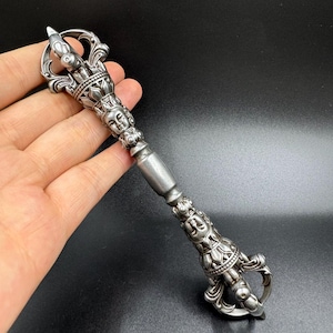 Puede incluir: Un vajra plateado y ornamentado, un objeto ritual con detalles intrincados. El vajra presenta un mango central y diseños elaborados, incluyendo caras y patrones florales. El objeto se sostiene en una mano sobre un fondo oscuro.