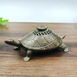 Puede incluir: Un quemador de incienso con forma de tortuga de color bronce. La tortuga tiene un caparazón detallado con una parte superior perforada y una pequeña tapa. El quemador está sobre una superficie de madera.