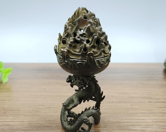 May include: A bronze-colored incense burner with a dragon base. The top section has a textured, flame-like design with multiple openings. The dragon is intricately detailed, with scales and a curved body. The burner is designed to hold incense.