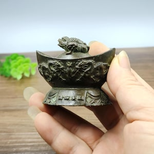 May include: A bronze-colored, decorative bowl with a frog figurine on top. The bowl has intricate dragon and floral patterns. The base of the bowl has engraved text. The item is held in a hand.