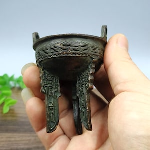 May include: A small, bronze-coloured, three-legged vessel with two handles. The vessel features intricate, embossed patterns around the rim and on the legs. The antique-style piece is held in a hand.