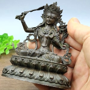 Könnte beinhalten: Bronzestatue einer sitzenden Gottheit, geschmückt mit einer Krone und aufwendigem Schmuck. Die Figur hält einen gebogenen Gegenstand in einer Hand und ruht auf einem Lotusblüten-Sockel. Die Statue wird in einer Hand gehalten.