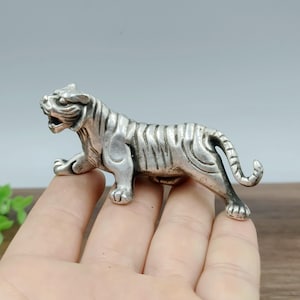 May include: A silver-colored tiger figurine, detailed with stripes and a snarling expression. The small sculpture is posed in a crouching position, showcasing its paws and tail. The figurine is made of metal and has a polished finish.