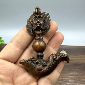 May include: Bronze-coloured religious artifact depicting a deity's face with a crown and flowing hair. The artifact has a bulbous midsection and a curved, ornate base. A small, clear stone is set at the end of the base.