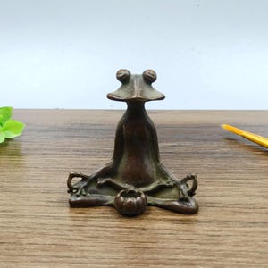 Puede incluir: Una figura de rana de bronce en pose de yoga, con las piernas cruzadas y las manos apoyadas en las rodillas. La rana tiene ojos grandes y redondos y una expresión serena. Un pequeño objeto en forma de calabaza está frente a la rana. La figura está sobre una superficie de madera.