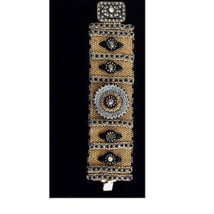 May include: A beaded bracelet with a rectangular clasp. The bracelet features a tan and black pattern with decorative elements, including a central circular design and diamond-shaped accents. The clasp is silver-toned and adorned with a central jewel.