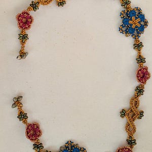 Puede incluir: Un collar de cuentas con un diseño floral. El collar tiene cuentas de color dorado con detalles en azul, rojo y verde. Los elementos florales están agrupados a lo largo del collar, creando un patrón decorativo. El collar se muestra sobre una superficie blanca.
