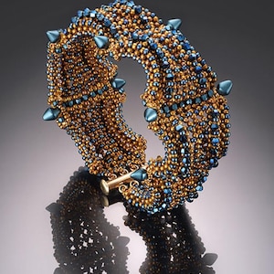 May include: A beaded bracelet with gold and blue beads. The bracelet has a gold clasp and is adorned with blue, teardrop-shaped accents. The bracelet is displayed on a reflective surface.