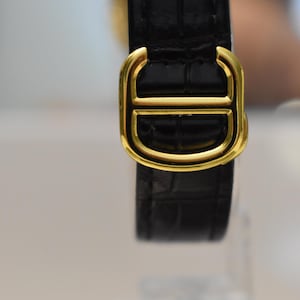 May include: Close-up of a black leather watch strap with a gold-colored buckle. The buckle has a distinctive design with a central bar and a D-shaped outline. The leather has a textured, crocodile-like pattern.