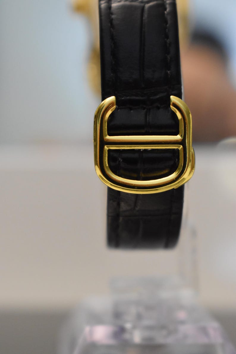 May include: Close-up of a black leather watch strap with a gold-colored buckle. The buckle has a distinctive design with a central bar and a D-shaped outline. The leather has a textured, crocodile-like pattern.