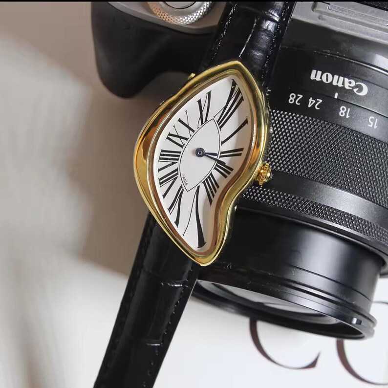 May include: A gold-toned wristwatch with a white face and black Roman numerals. The watch has a unique, melting design and a black leather band. The watch face reads "Paris".