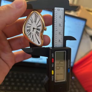 May include: A gold-toned, melting-style wristwatch with a white face, black Roman numerals, and black hands. The watch is being measured with a digital caliper, showing a measurement of 46.1 mm.