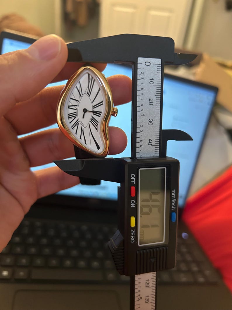 May include: A gold-toned, melting-style wristwatch with a white face, black Roman numerals, and black hands. The watch is being measured with a digital caliper, showing a measurement of 46.1 mm.