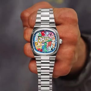 May include: A stainless steel wristwatch with a colorful, artistic dial featuring various cartoon figures and shapes. The watch has a date display at the 3 o'clock position and a silver band. The watch is held by a person.