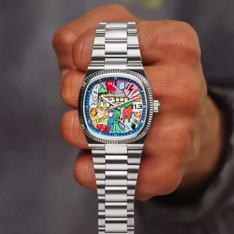 May include: A stainless steel wristwatch with a colorful, artistic dial featuring various cartoon figures and shapes. The watch has a date display at the 3 o'clock position and a silver band. The watch is held by a person.