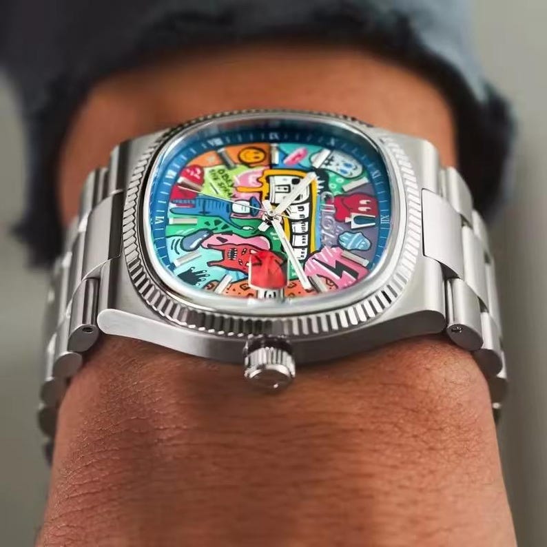 May include: A stainless steel wristwatch with a colorful, graffiti-style dial. The watch features a silver band and a crown on the side. The dial has a blue background with various cartoon-like images and the word "FREE" in white.