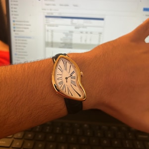 May include: A gold-toned wristwatch with a white face and black Roman numerals. The watch has a unique, melting design and a black leather band. The watch is worn on a wrist.