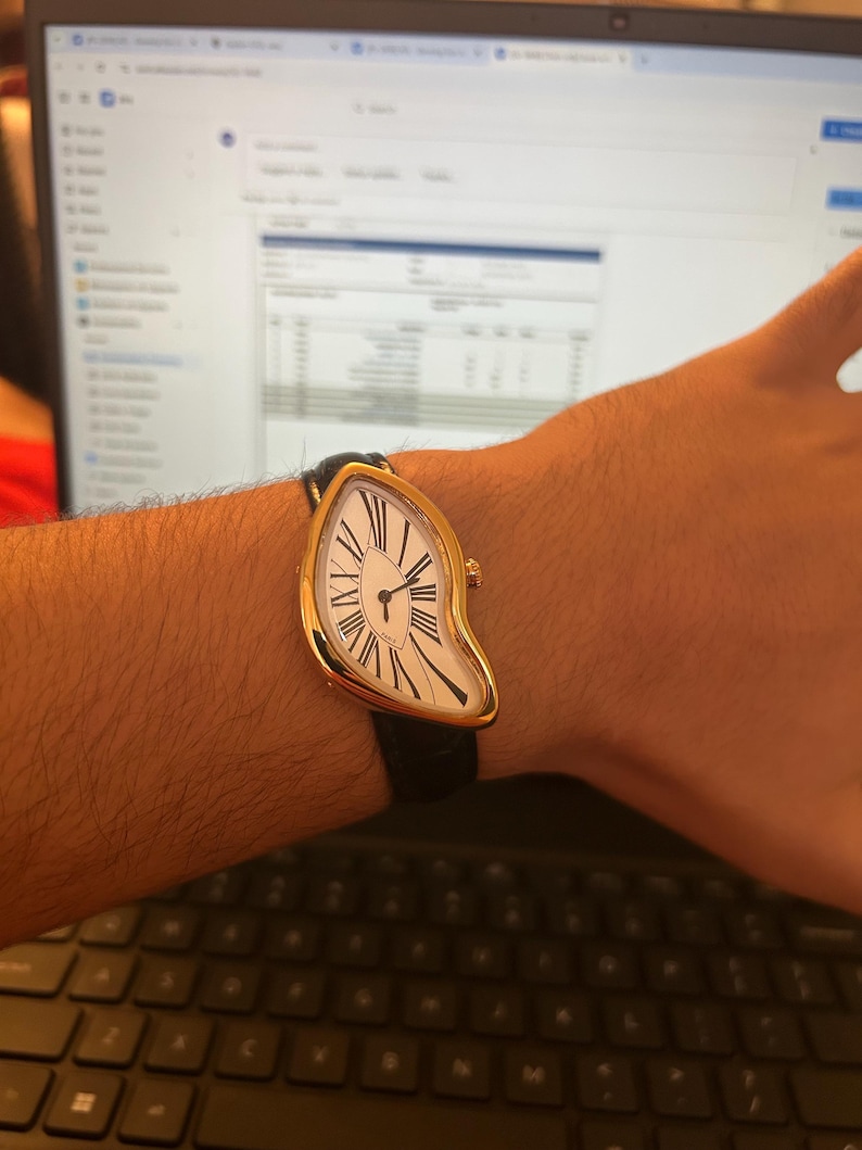 May include: A gold-toned wristwatch with a white face and black Roman numerals. The watch has a unique, melting design and a black leather band. The watch is worn on a wrist.