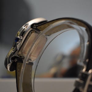 May include: Close-up of a silver wristwatch with a rectangular face and a blue gem. The watch has a black leather band and a clear, curved protective cover. The watch is a luxury item.