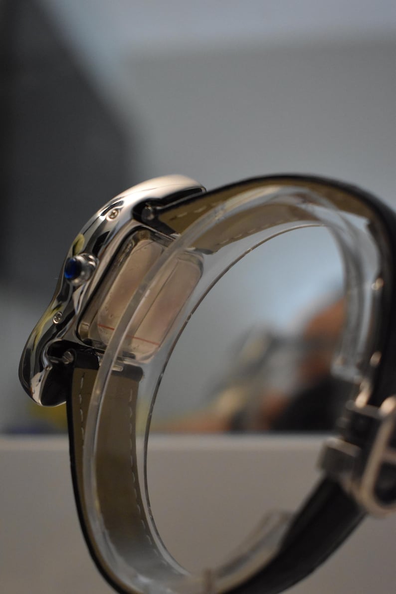 May include: Close-up of a silver wristwatch with a rectangular face and a blue gem. The watch has a black leather band and a clear, curved protective cover. The watch is a luxury item.