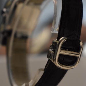 May include: Close-up of a luxury wristwatch with a black leather band and a silver-colored buckle. The watch is displayed on a clear stand, showcasing the band's texture and the buckle's design. The watch face is out of focus.