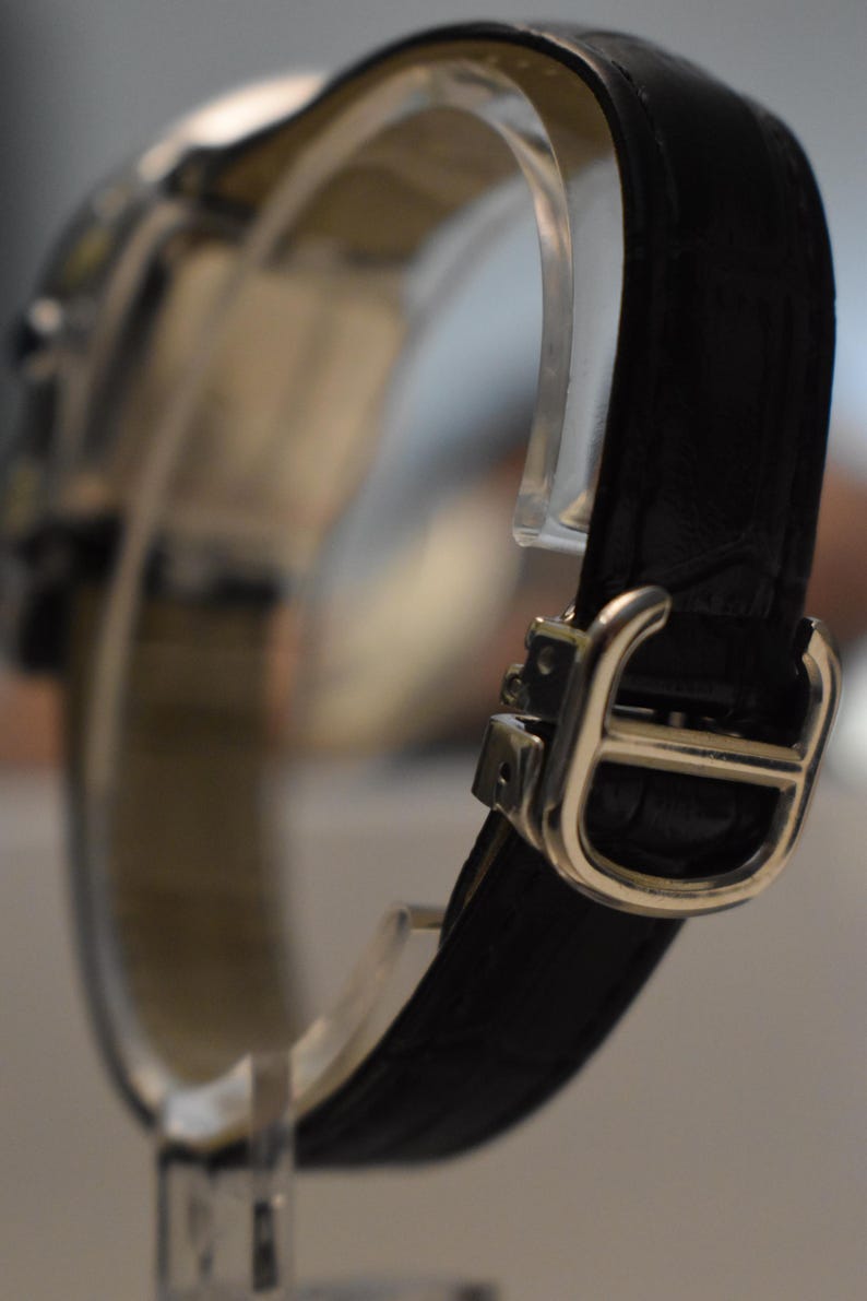 May include: Close-up of a luxury wristwatch with a black leather band and a silver-colored buckle. The watch is displayed on a clear stand, showcasing the band's texture and the buckle's design. The watch face is out of focus.