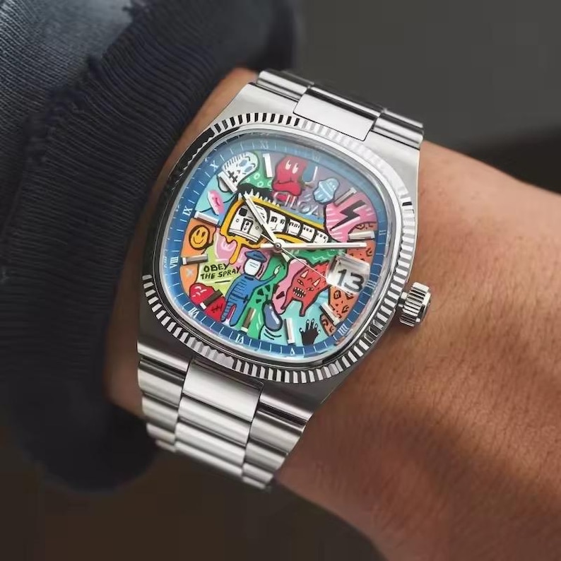 May include: A stainless steel wristwatch with a colorful, graffiti-style face. The watch features a square case, a fluted bezel, and a date display at the 3 o'clock position. The dial is adorned with cartoon characters and the text "OBEY THE SPRAY".