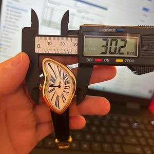 May include: A gold-toned wristwatch with a white face and black Roman numerals. The watch has a unique, curved shape and a black leather band. The digital caliper shows a measurement of 30.2 mm.
